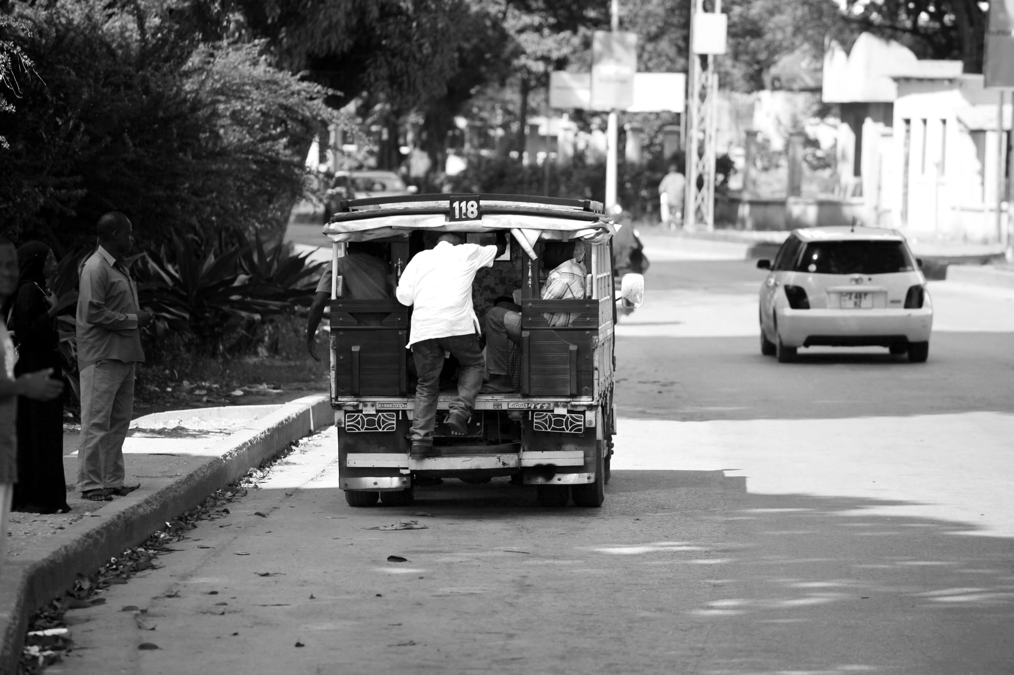 A black and white street photograph taken in Zanzibar, capturing the rear of a moving dala dala, a traditional open-air minibus. A passenger in a white shirt is climbing into the crowded back of the vehicle, which is marked with the number 118 on its roof. The dala dala travels on a sunlit paved road, flanked by leafy trees and buildings. Several pedestrians wait on the left sidewalk, while a light-colored hatchback car drives in the distance ahead of the bus.