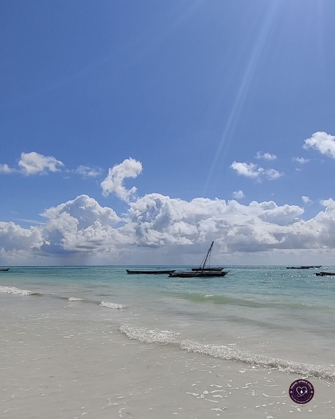 Vertical shot of Jambiani Beach in Zanzibar under a bright blue sky filled with dramatic white cumulus clouds and a sun flare ray streaming down. Shallow, clear turquoise waves gently lap onto soft white sand in the foreground, while traditional wooden dhow boats rest anchored in the calm open water further out. A small circular logo for "Travel Africa Solo" is in the bottom right corner.