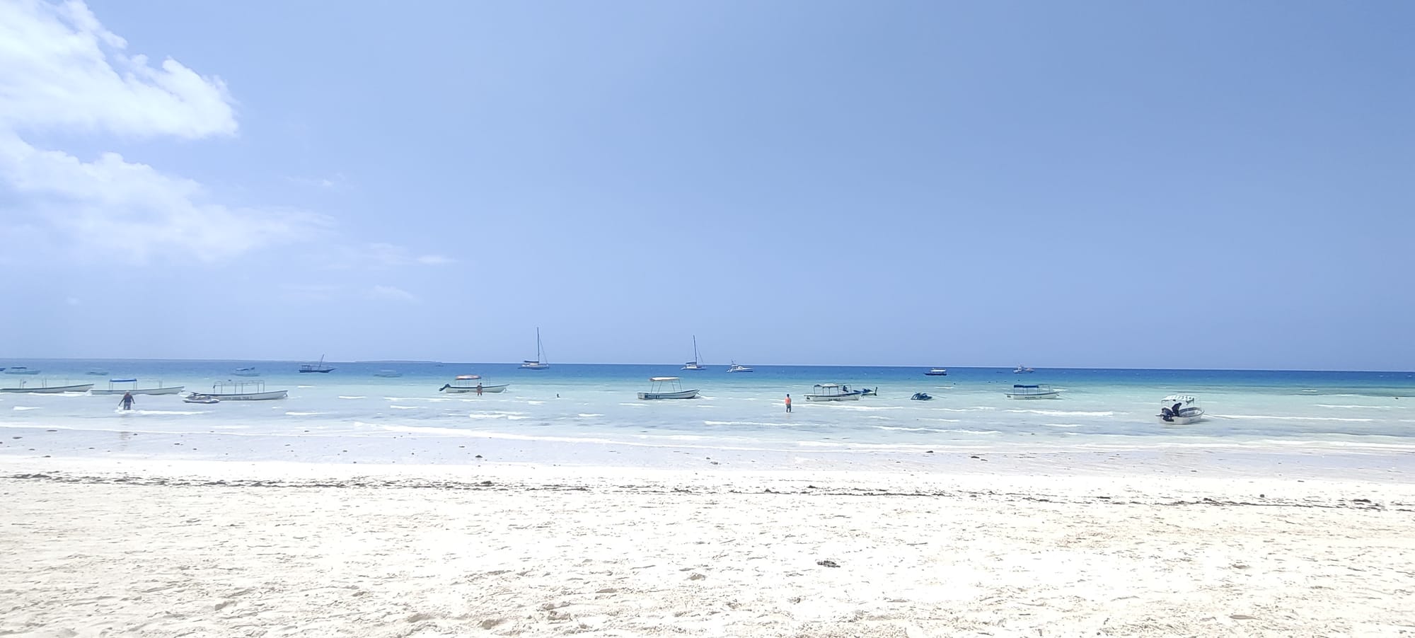 Wide-angle view of Nungwi Beach in Zanzibar featuring powdery white sand in the foreground and calm, crystal-clear turquoise ocean waters. Several small boats and traditional dhows float anchored along the horizon under a bright, sunny blue sky with scattered light clouds.